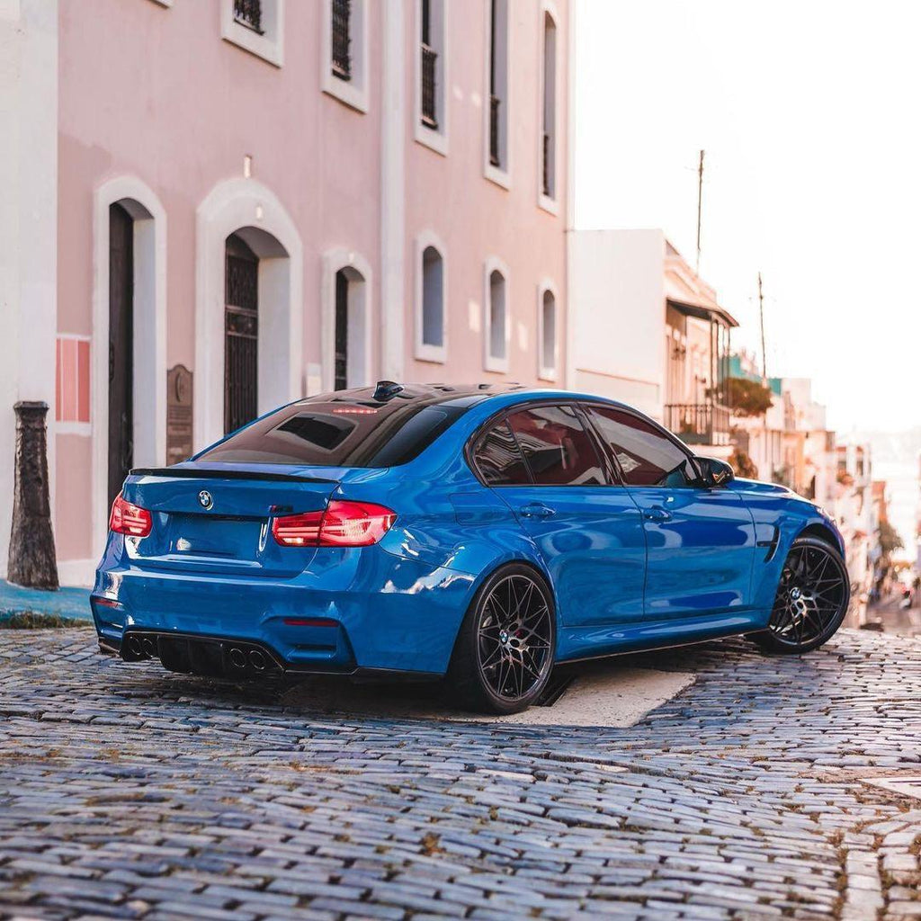 Kék BMW F80 M3 ferdén hátulról macskaköves utcán, carbon csomagtartó spoilerrel (MP Style)