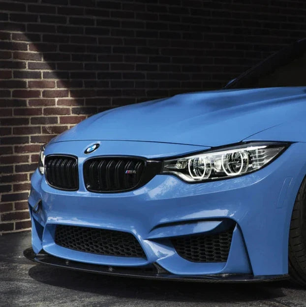 Blue BMW M3/M4 front close-up in front of a brick wall, with carbon fiber front lip (3D Style)