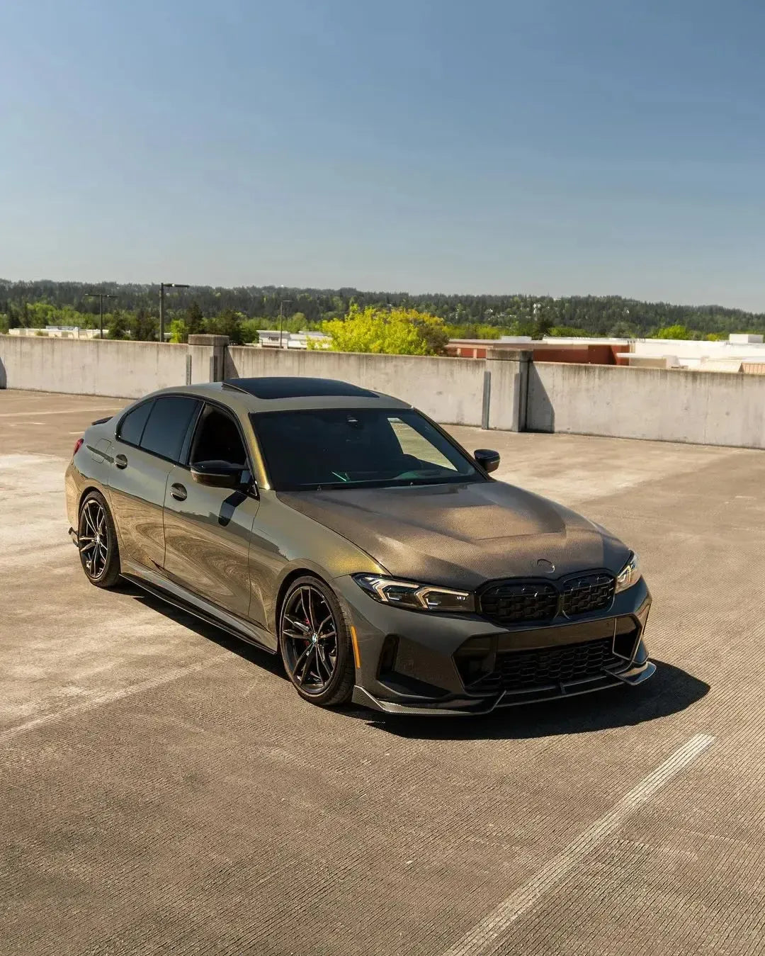 Matte olive gray BMW G20 LCI photographed on a rooftop, front three-quarter view, with carbon side skirts (CMST Style)