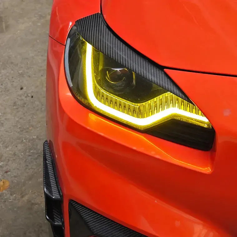 Narancssárga BMW G87 M2 fényszórója közelről, felszerelt carbon fényszóró szemöldökkel