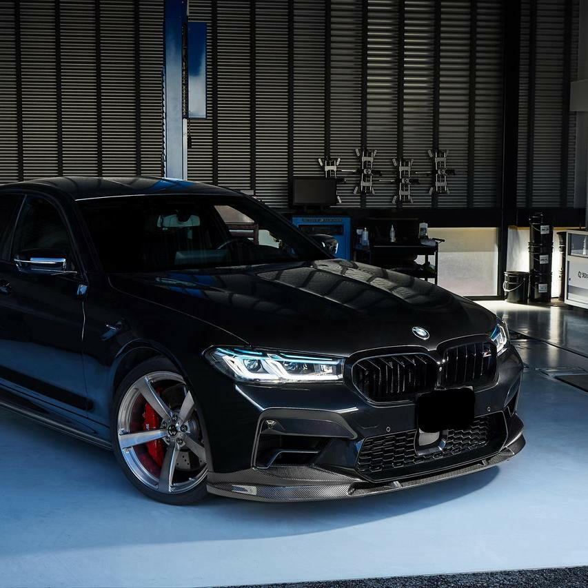 Black BMW F90 M5 LCI in a workshop, front three-quarter view, with carbon front lip (3D Style)