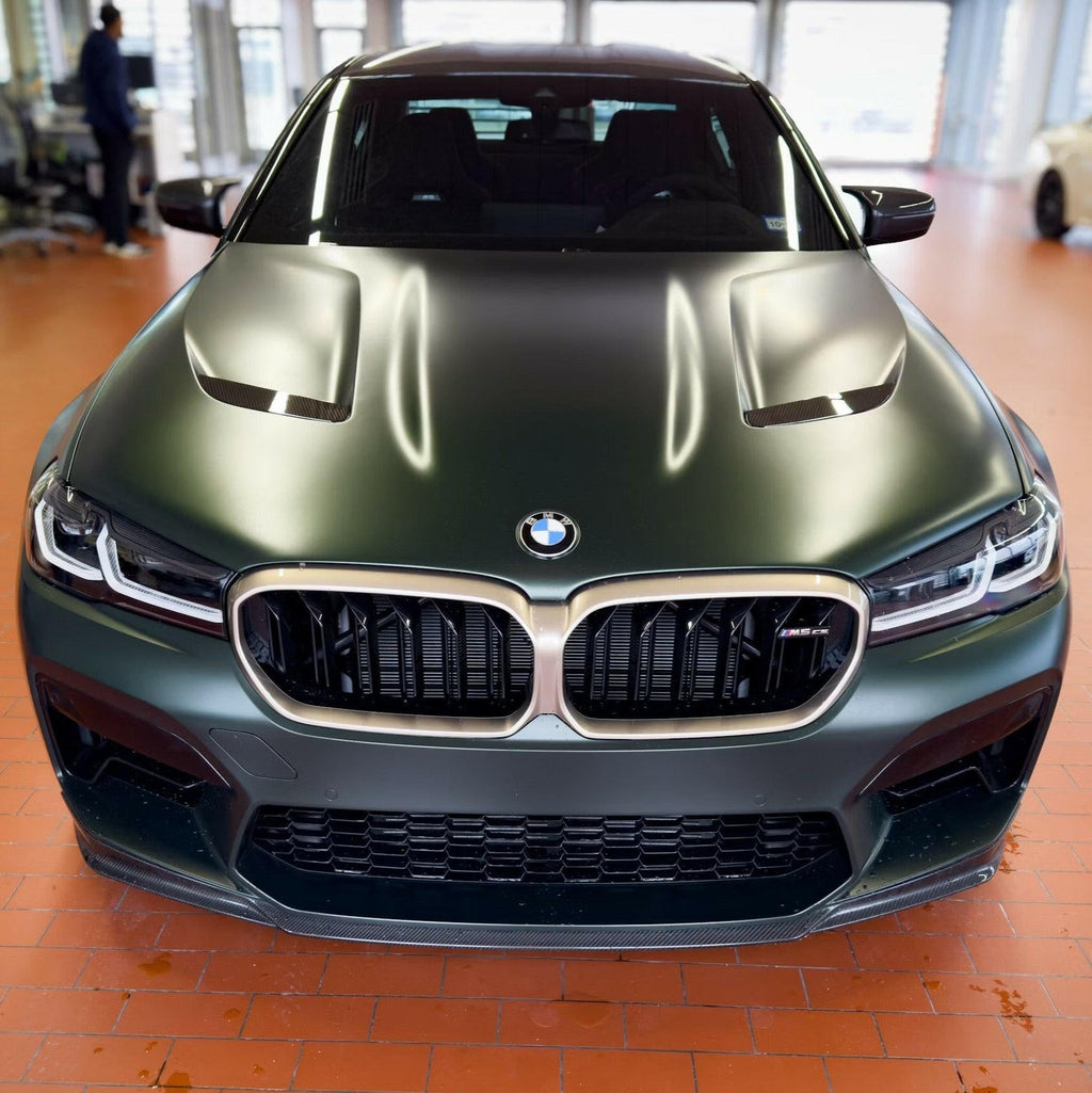 Matte green BMW F90 M5 CS LCI head-on in a showroom, with carbon front lip and vented hood (CS Style)