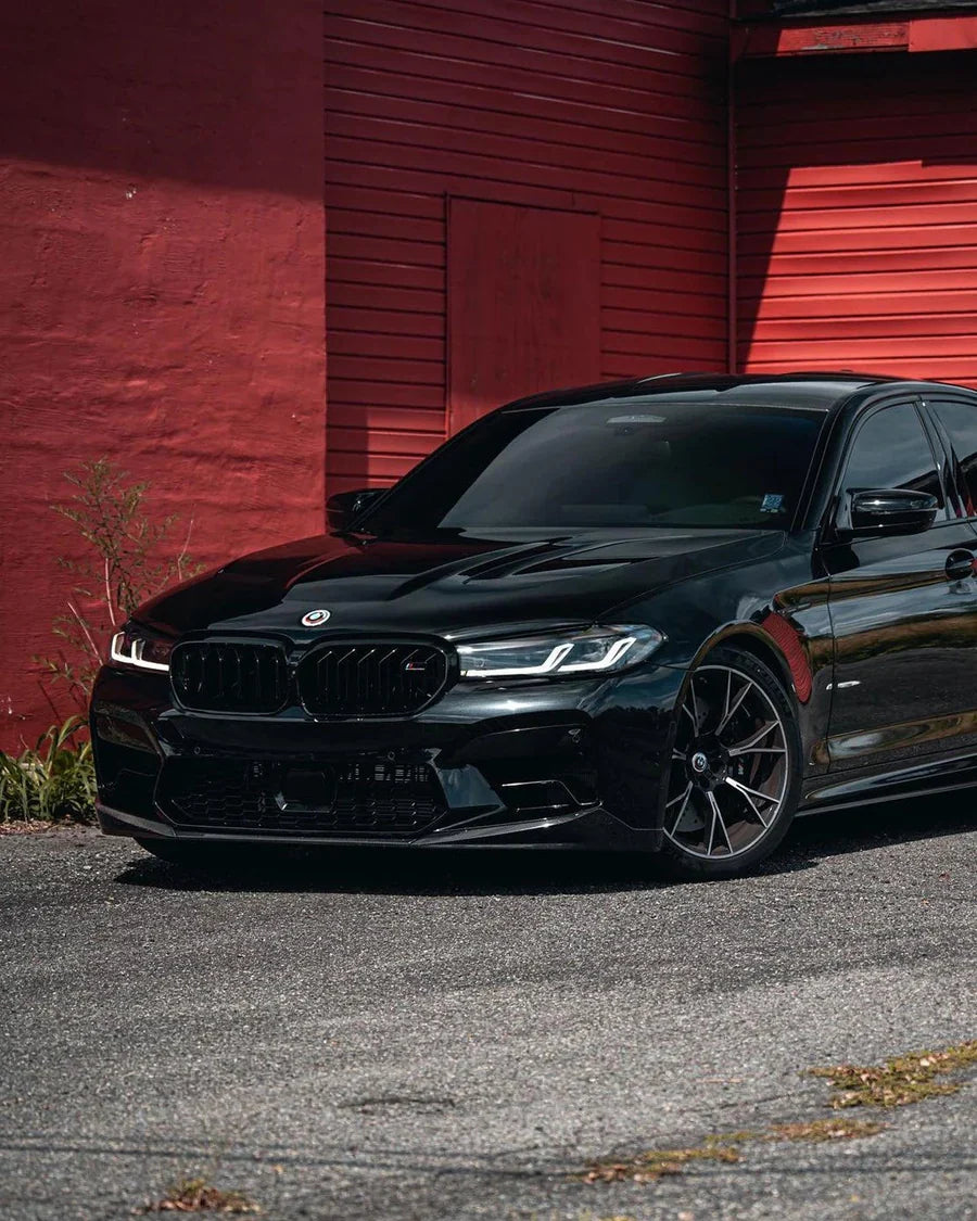 Black BMW F90 M5 LCI in front of a red wall, front three-quarter view, with fitted carbon front lip (PM V1 Style)