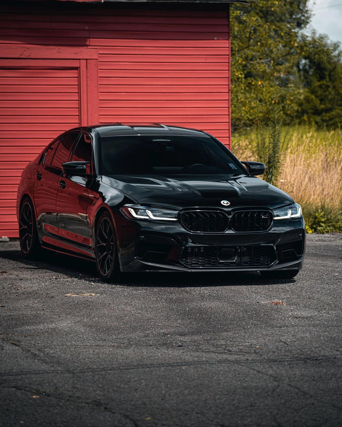 Black BMW F90 M5 LCI in front of a red barn, front three-quarter view, with carbon front lip (PM V1 Style)