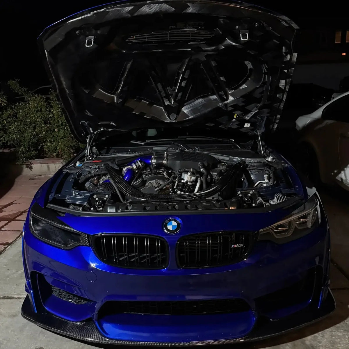 Kék BMW F80 M3 szemből éjjel, nyitott motorháztetővel és fényszóró helyetti carbon légbeömlővel