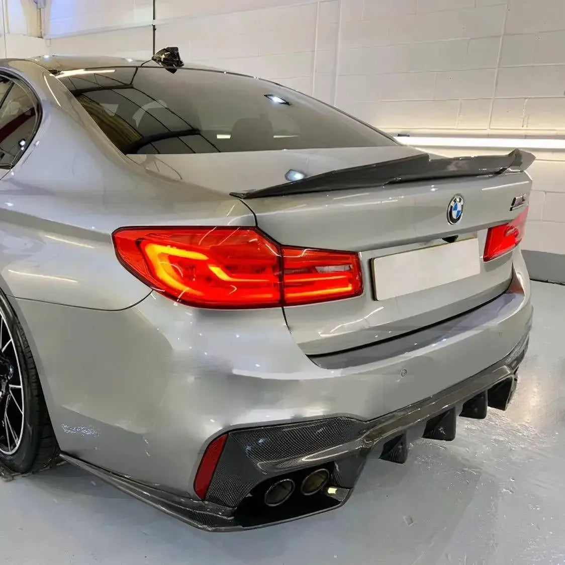 Silver pre-LCI BMW F90 M5 in rear three-quarter view in a garage, with carbon spoiler (PSM Style High Kick)